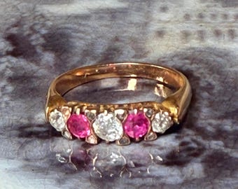 May include: A gold ring featuring a series of alternating gemstones. The ring has a warm, golden hue, with bright white and vibrant pink stones set across the top. The gemstones are round and set close together.
