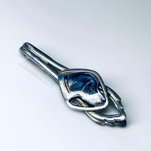 May include: Silver-toned tie clip featuring a rectangular design with a painted dog portrait in blue, white, and black enamel. The clip has a curved, elongated shape with a loop at the end.