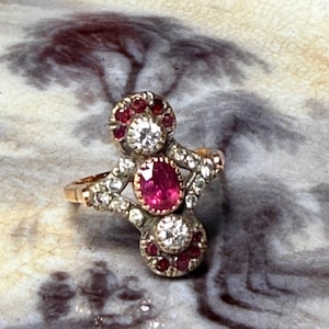 May include: An antique gold ring with a central oval ruby, encircled by smaller rubies and diamonds. The ring has an intricate, ornate design with a vintage aesthetic. The ring's setting is a mix of gold and silver.