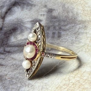 May include: Vintage gold ring with a marquise-shaped face. The ring features a large central pearl surrounded by a halo of small red stones, flanked by two smaller pearls. The ring's setting is detailed with small diamonds and filigree.