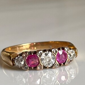 Peut inclure: Une bague en or avec cinq pierres serties en rangée. Les pierres alternent rose et blanc. La bague est sur une surface blanche.