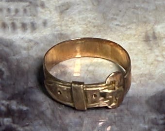 May include: A gold-colored ring with a buckle design. The ring has a band that resembles a belt, complete with a rectangular buckle and a belt loop. The ring is made of a shiny, metallic material.