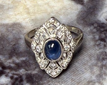 May include: A silver ring featuring an oval, deep blue gemstone at the center, surrounded by a decorative, ornate setting with small, clear accent stones. The ring has a vintage or antique aesthetic.