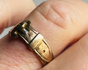 May include: A gold-coloured ring in the shape of a belt buckle. The ring features a detailed design with a buckle and belt loops. The ring is worn on a finger.