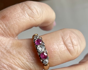 May include: A gold ring featuring a row of alternating gemstones. The ring has two bright red rubies and two clear, sparkling diamonds, with smaller accent stones. The ring is held by a person's hand.