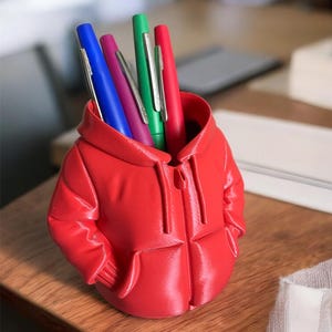 Peut inclure: Un porte-stylos rouge en forme de sweat à capuche, contenant des stylos bleus, violets, verts et rouges. Le porte-stylos a une capuche, des poches et une fermeture éclair. Le porte-stylos est sur une surface en bois.
