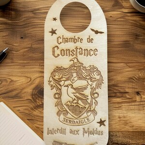 Peut inclure: Un porte-nom de porte en bois avec un design inspiré de Poudlard. Il présente un blason avec un oiseau, le texte "Serdaigle", et les mots "Chambre de Constance" et "Interdit aux Moldus".
