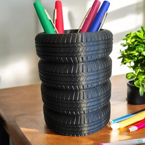 Peut inclure: Un pot à crayons noir fait de pneus empilés, rempli de stylos colorés. Le pot à crayons est posé sur une surface en bois, avec d'autres stylos éparpillés à proximité. Une petite plante verte est en arrière-plan.