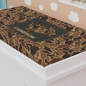 Puede incluir: Una funda de cambiador de color marrón y beige con camuflaje con el nombre "CAMERON" impreso en ella.