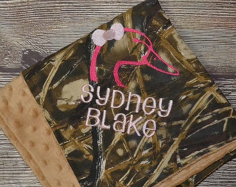 May include: A square baby blanket featuring a camouflage print on one side and a tan dimpled minky fabric on the other. The camouflage side is embroidered with a pink duck silhouette wearing a bow and the name SYDNEY BLAKE in pink lettering.