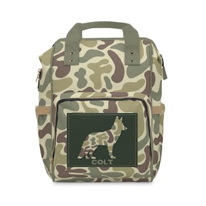 Puede incluir: Mochila de camuflaje con un parche verde que presenta la silueta de un perro pastor alemán y la palabra "COLT".