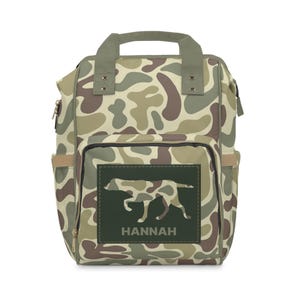 May include: A camouflage-patterned backpack with a green patch featuring a silhouette of a dog and the name "HANNAH".