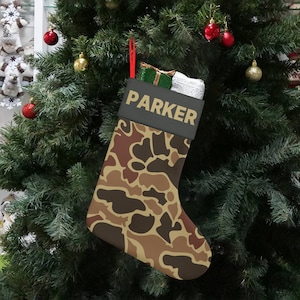 May include: A Christmas stocking with a camouflage pattern in shades of brown and tan hangs on a decorated Christmas tree. The stocking has a dark gray cuff with the name "PARKER" printed in gold. The stocking is filled with gifts.