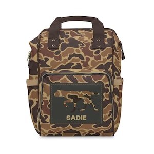 May include: A camouflage-patterned diaper bag backpack with a brown leather handle and a black patch with a dog silhouette and the name "SADIE" in white text.