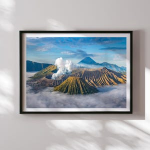 Pode incluir: Impressão emoldurada de uma paisagem vulcânica. A imagem mostra vários picos vulcânicos, alguns com crateras visíveis e colunas de fumaça, contra um fundo de céu azul e nuvens. O primeiro plano é envolto por uma camada de névoa.