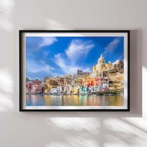 May include: A colourful cityscape with a blue sky and white clouds. The buildings are clustered together on a hillside and have a variety of colours, including yellow, orange, red, and green. There is a body of water in the foreground, and the reflection of the buildings can be seen in the water.