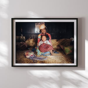 May include: An older woman wearing a straw hat and a colorful patterned dress sits on a woven mat with a young child on her lap. The child is wearing a red and black plaid dress and a necklace of beads. The woman and child are smiling at the camera.