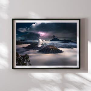 Op de afbeelding: Een ingelijste landschapsfoto van een vulkanisch berglandschap gehuld in mist. De afbeelding toont een dramatische lucht met blikseminslagen, wat een dynamisch element aan de scène toevoegt. De voorgrond toont de toppen van de vulkanen.
