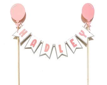 May include: Pink and white cake topper with the name "HADLEY" in pink letters on a white banner with silver trim. The banner is attached to two wooden sticks with pink balloons on top.
