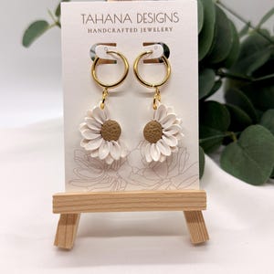 Könnte beinhalten: Ohrringe mit Gänseblümchen-Design, goldenen Reifen und einem braunen Mittelteil. Die Ohrringe werden auf einer kleinen Holzstaffelei mit dem Text "TAHANA DESIGNS HANDCRAFTED JEWELRY" auf der Karte dahinter präsentiert. Der Hintergrund ist weiß mit grünem Laub.