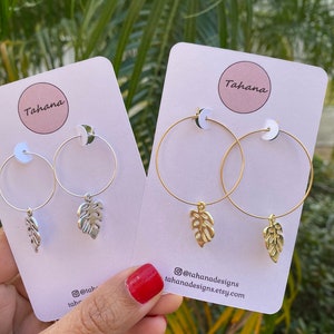 May include: Two pairs of hoop earrings with a leaf charm. The earrings are on a white card with the word "Tahana" and the social media handle "@tahanadesigns" printed on it.