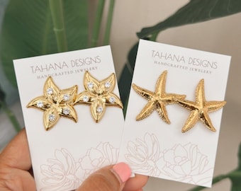 Puede incluir: Dos pares de pendientes de estrella de mar dorados sobre tarjetas blancas. Un par tiene detalles de cristal transparente. Las tarjetas dicen "TAHANA DESIGNS HANDCRAFTED JEWELRY". Los pendientes se muestran sobre un fondo de follaje verde.