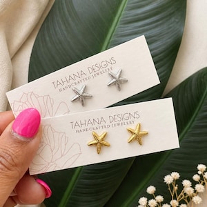 Puede incluir: Dos pares de pendientes de botón con forma de estrella de mar, uno plateado y otro dorado, presentados en tarjetas con el texto "TAHANA DESIGNS HANDCRAFTED JEWELRY". Los pendientes están sobre hojas verdes y flores blancas.
