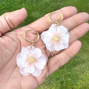以下が含まれることがあります： 白い花のチャームが付いたゴールドのフープピアスのペア。花は繊細で透き通った外観で、小さなゴールドのセンターがあります。