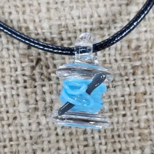 May include: A glass pendant necklace with a blue glass spool of thread and a black glass needle. The pendant is on a black cord.