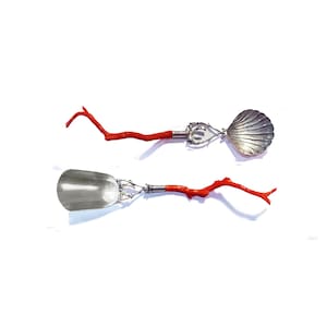 May include: A set of two silver serving spoons with red coral handles. The spoons are shaped like a seashell and a scoop.