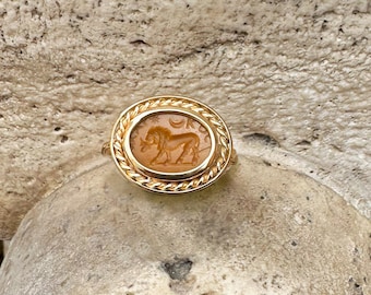 May include: A gold ring with an oval amber-coloured stone, carved with a lion design. The ring has a braided rope-like border and a simple gold band. The ring is set against a neutral, textured background.