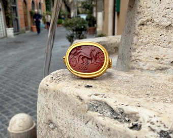May include: A gold ring with an oval, red stone featuring a carved design of a horse-drawn chariot. The ring has a gold band and is set on a stone surface. The ring is a piece of jewellery.