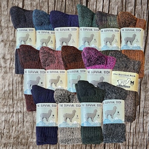 Puede incluir: Una variedad de calcetines de lana de alpaca en diferentes colores y tamaños. Los calcetines están hechos en los EE. UU. con lana de alpaca cultivada en los EE. UU. Los calcetines están etiquetados con el texto "The Survival Sock" y "Heavyweight Full Cushion Crew".