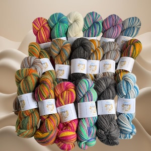 Puede incluir: Una colección de madejas de hilo coloridas en varios tonos, incluyendo rosa, verde, amarillo y azul. Cada madeja está envuelta con una etiqueta blanca. El hilo está dispuesto en una exhibición visualmente atractiva, ideal para proyectos de tejido o ganchillo.