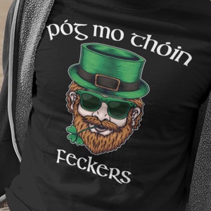 May include: Black t-shirt with a cartoon illustration of a leprechaun wearing a green hat and sunglasses. The text "póg mo chóin" and "Feckers" is printed on the shirt.