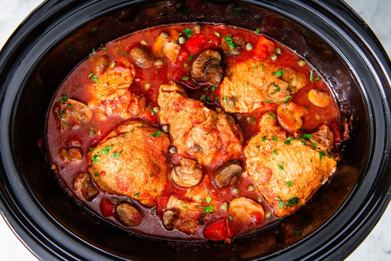 May include: A close-up of a slow cooker filled with chicken, mushrooms, peppers, and a red sauce. The chicken is cooked and the sauce is bubbling.
