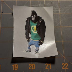 May include: A sticker featuring a cartoon gorilla character from the movie "Sing". The gorilla is wearing a black leather jacket, a green shirt with a yellow lightning bolt, blue jeans, and white sneakers.