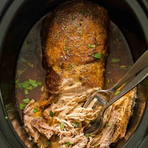 May include: A close-up of a slow cooker filled with shredded pork, with a spoon in the pot. The pork is golden brown and has a crispy exterior. The pork is surrounded by a flavorful broth.