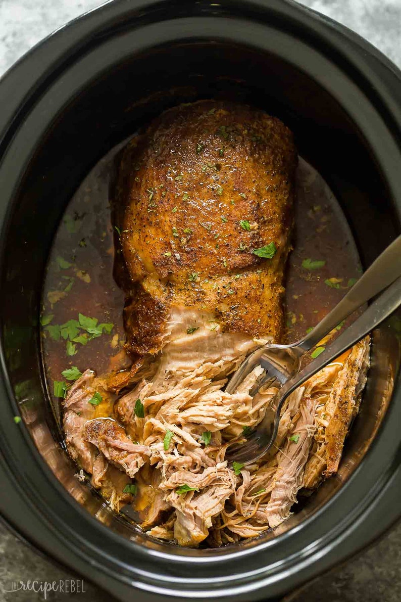 May include: A close-up of a slow cooker filled with shredded pork, with a spoon in the pot. The pork is golden brown and has a crispy exterior. The pork is surrounded by a flavorful broth.