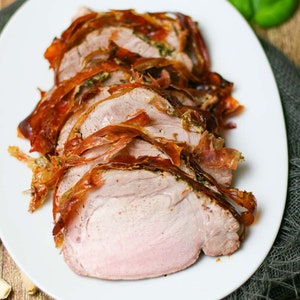 May include: Close-up of a sliced pork loin wrapped in prosciutto and roasted. The pork is on a white plate with a gray napkin underneath.