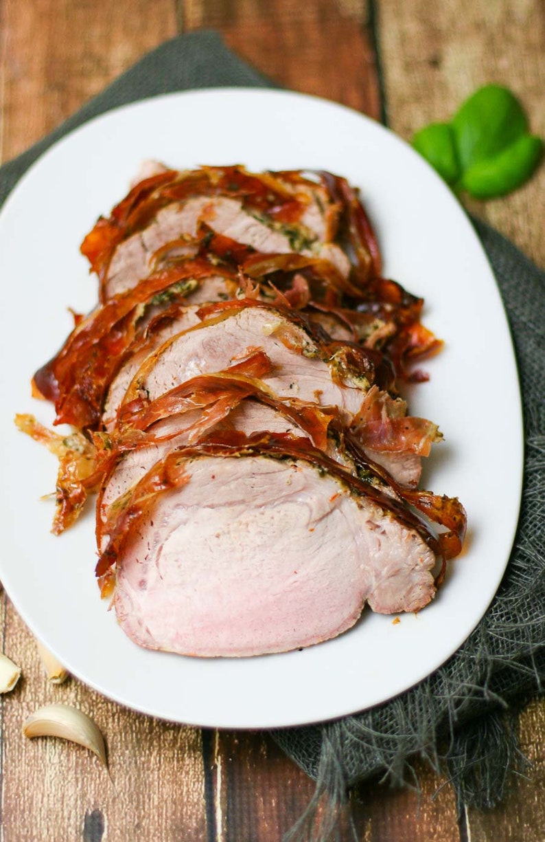 May include: Close-up of a sliced pork loin wrapped in prosciutto and roasted. The pork is on a white plate with a gray napkin underneath.
