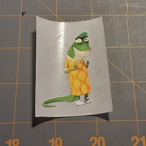 May include: A green lizard wearing a yellow dress and white shoes. The lizard has a surprised expression on its face.