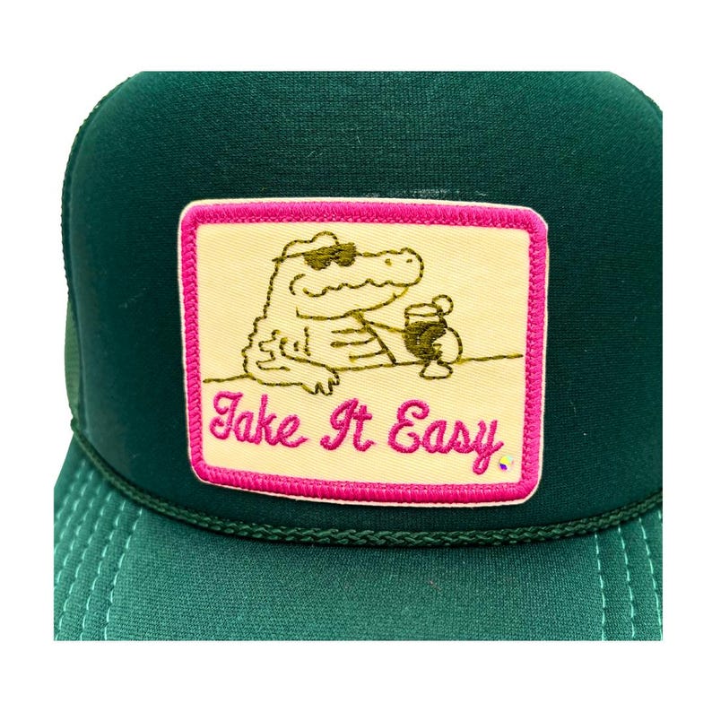Pode incluir: Bon&eacute; de caminhoneiro verde com um patch com um jacar&eacute; usando &oacute;culos de sol e segurando uma bebida. O patch tem o texto "Take It Easy" em rosa. O bon&eacute; tem uma parte traseira de malha e uma aba curva.