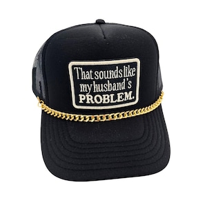 May include: Black trucker hat with a mesh back and a curved brim. The front features a rectangular patch with the embroidered text "That sounds like my husband's PROBLEM." A gold-colored chain is attached to the brim.