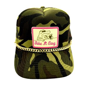 Pode incluir: Bon&eacute; de caminhoneiro camuflado com corrente dourada e um patch com um jacar&eacute; usando &oacute;culos de sol e segurando uma bebida. O patch tem o texto "Take It Easy" em rosa e branco.