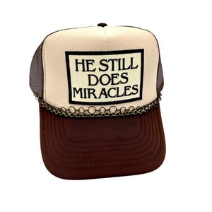 May include: A brown and tan trucker hat with the text "HE STILL DOES MIRACLES" in a black rectangle. The hat has a brown brim, a tan front panel, and a mesh back. A decorative chain is attached to the brim.