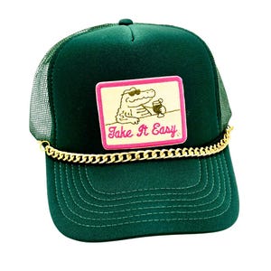 Pode incluir: Bon&eacute; de caminhoneiro verde escuro com parte traseira em rede e aba curva. A frente apresenta um patch com um jacar&eacute; usando &oacute;culos de sol e segurando uma bebida, com o texto "Take It Easy". Uma corrente dourada adorna a aba.
