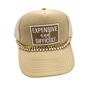May include: Tan trucker hat with a mesh back and a curved brim. The hat features a patch with the words "EXPENSIVE and DIFFICULT" in white and gold lettering. A gold chain with faux pearls is attached to the brim.