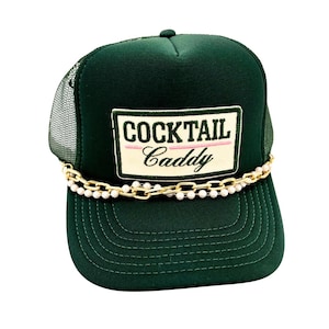 May include: A dark green trucker hat with a mesh back and a curved brim. The hat features a rectangular patch with the words "COCKTAIL CADDY" in white and pink lettering. A gold chain with white beads is attached to the front.