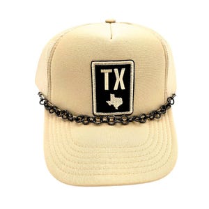 May include: Tan trucker hat with a mesh back and a curved brim. The front features a black and white patch with the letters "TX" and a Texas outline. A black chain detail is attached to the brim.